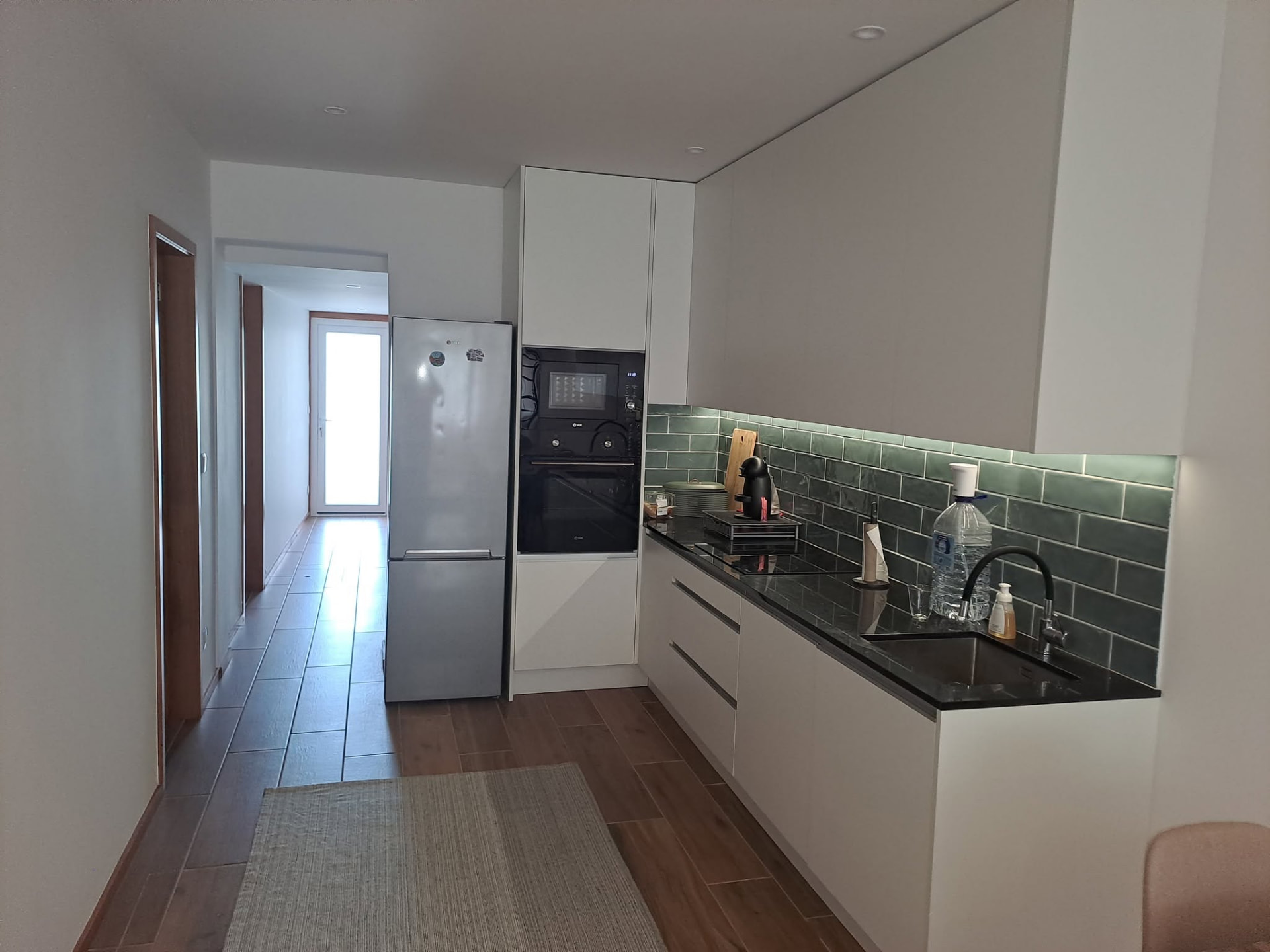Cozinha remodelada em open space com armários brancos, bancada escura e azulejo verde
