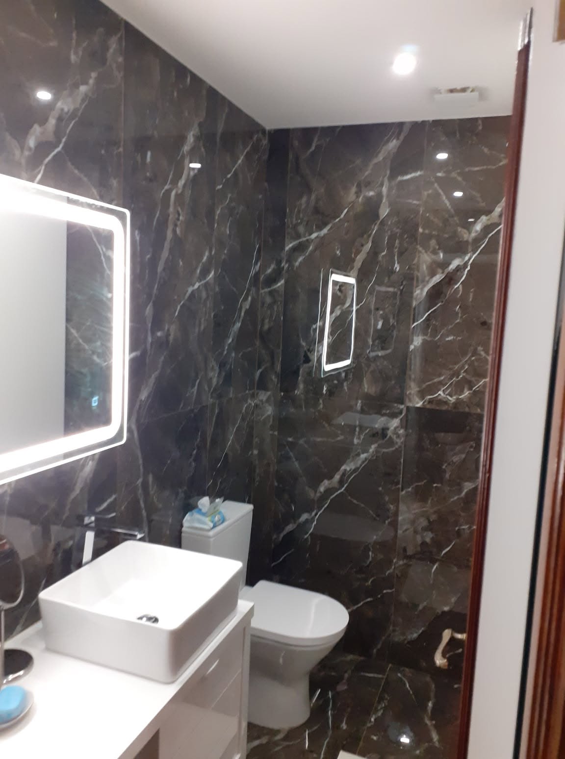 Casa de banho remodelada com revestimento escuro, lavatório branco e espelho retroiluminado